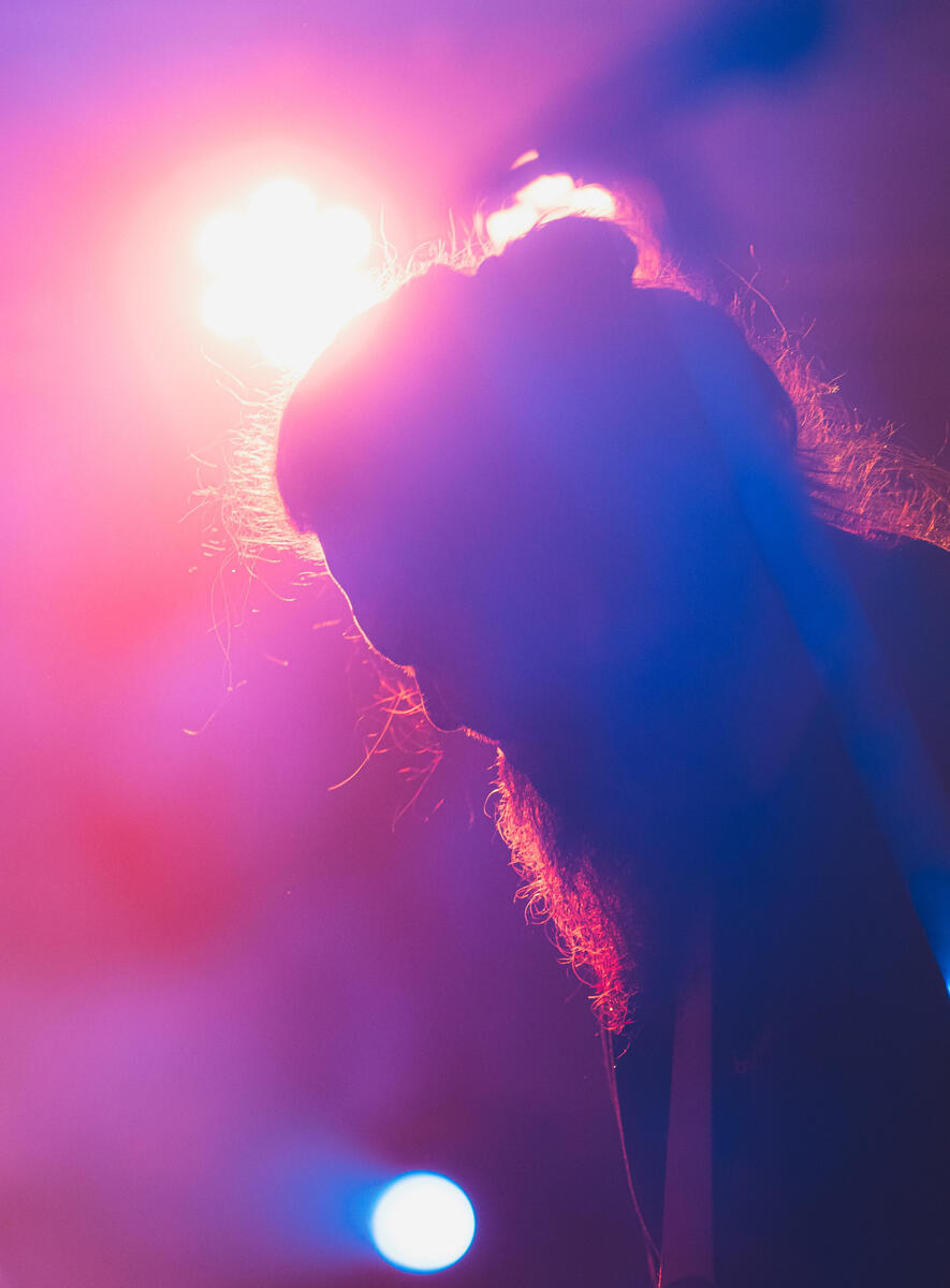 Close up aesthetic colour photo of guitarist's face and hair with red, pink, orange, blue, purple background light